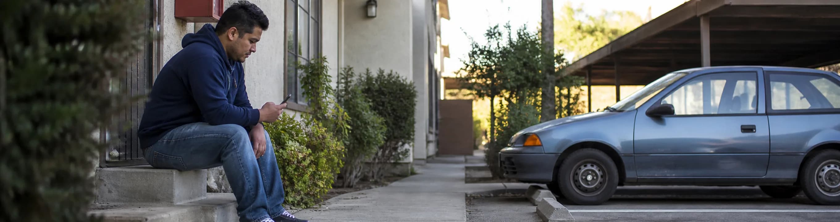 A person sitting on a front step outside a house, an older car parked under a carport nearby