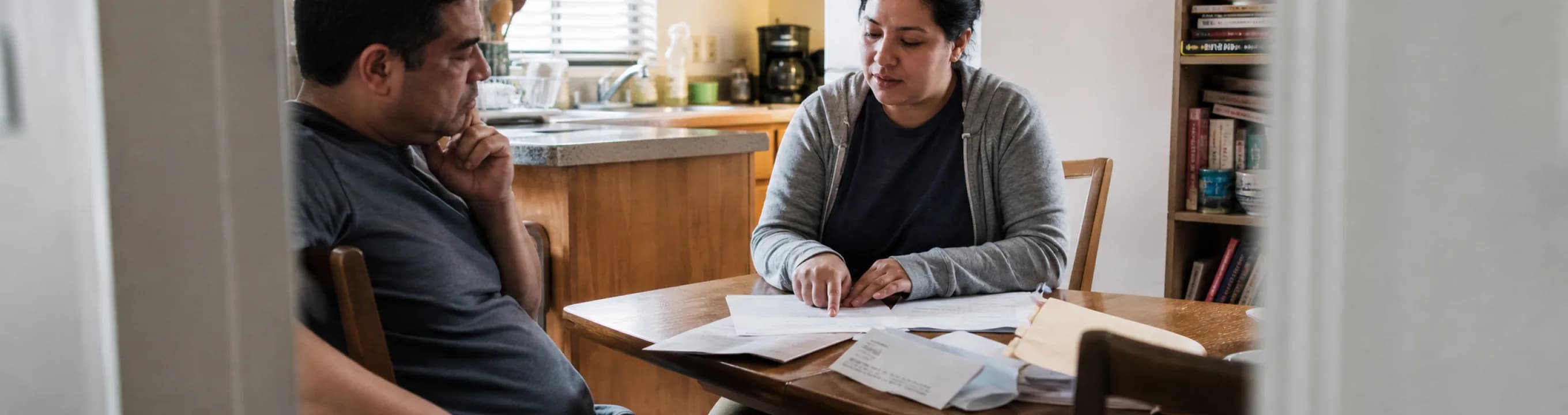 Two people at a kitchen table with statements and a written list spread between them