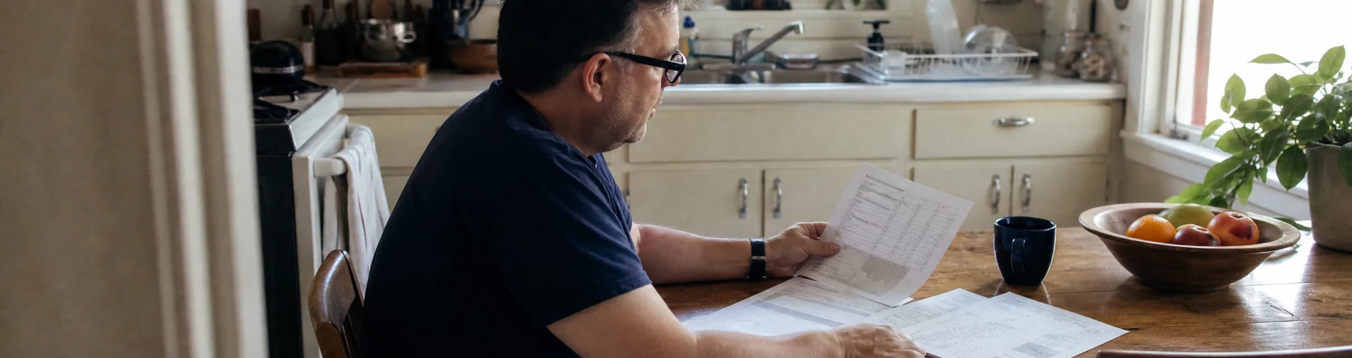 A person reading printed statements at a kitchen table beside a window